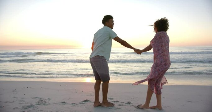 Biracial couple holds hands on a beach at sunset, waves gently breaking in the background