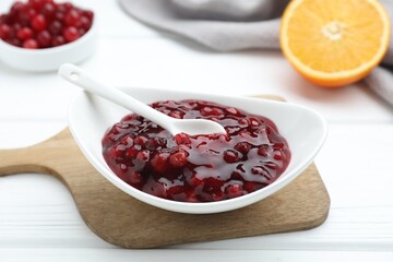 Fresh cranberry sauce in bowl and spoon on white wooden table, closeup