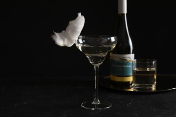 Cocktail with tasty cotton candy and bottle of alcohol drink on dark textured table against black background, space for text