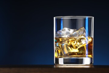 Whiskey with ice cubes in glass on table against blue background, closeup. Space for text