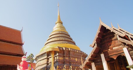 Golden pagoda wrapped in a saffron cloth against a clear blue sky, flanked by traditional Thai wooden structures, symbolizing cultural heritage at Wat Phra That Lampang Luang, northern Thailand
