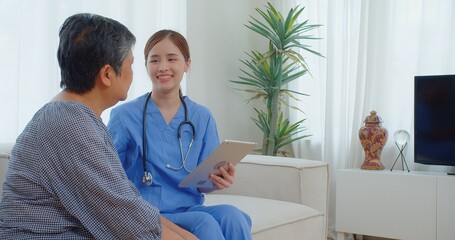 A healthcare professional in blue scrubs consults with an elderly patient using a tablet in a bright airy room