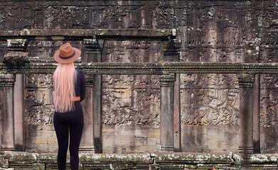 Fototapeta premium A beautiful blonde woman with a stylish hat wearing black tight trousers watching Bas-relief - Bas-relief at Bayon temple in Angkor Thom. Siem Reap. Cambodia. Panorama