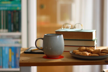 Cup of tea or coffee, pile of books, plate of cookies, reading glasses, e-reader and pen on the table. Colorful rainbow bookshelf in the background. Selective focus.