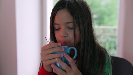 Little girl drinking warm drink during cold day indoors. Child sipping hot choco to stay warm during winter season