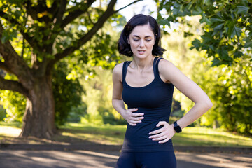 A young woman is running and doing sports in the park, standing with her hands on her stomach, feeling nauseous and near after exercise