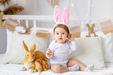 baby girl in bunny ears on the bed at home with painted eggs and rabbit, easter concept, cute...