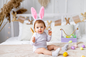 Fototapeta premium cute happy baby girl in bunny ears on the bed at home with a rabbit and painted eggs, easter concept, funny Easter baby is waiting for the holiday and rejoicing