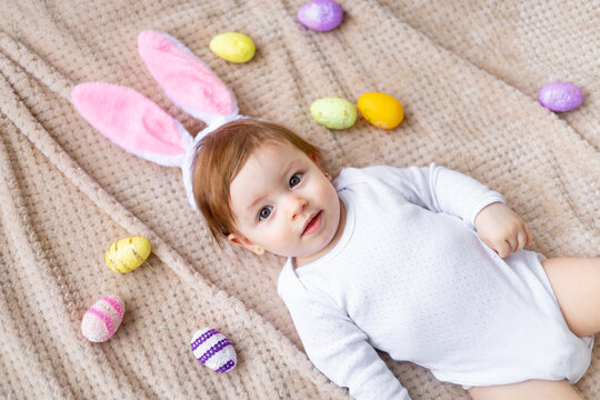 cute little baby with bunny ears and painted eggs lies on a beige background and smiles, easter concept