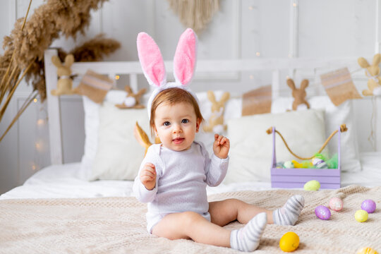 cute happy baby girl in bunny ears on the bed at home with a rabbit and painted eggs, easter concept, funny Easter baby is waiting for the holiday and rejoicing