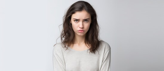 An attractive young brunette Caucasian woman with long hair looks unhappy, stressed, and depressed as she stands in front of a plain white wall. She appears isolated and alone. - obrazy, fototapety, plakaty