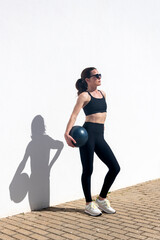 sporty woman standing by a white wall holding a pilates ball under her arm. oudoor fitness concept