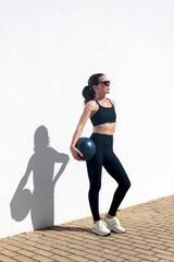 Obraz premium sporty woman standing by a white wall holding a pilates ball under her arm. oudoor fitness concept