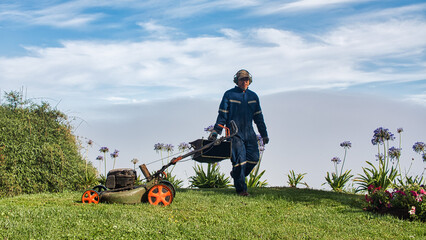 Yard worker mowing grass with lawn mower in garden. Lawn maintenance. - obrazy, fototapety, plakaty