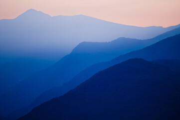 Stacked silhouettes of distant blue mountains underneath a homogenous orange evening sky at Lago...
