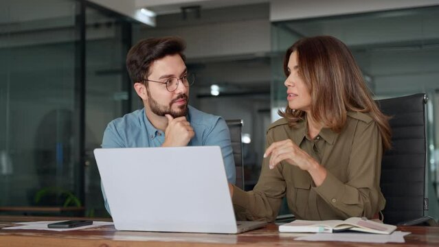 Two busy colleagues working together talking using laptop in office. Middle aged Latin female manager teaching young male worker looking at computer discussing business plan at office meeting.