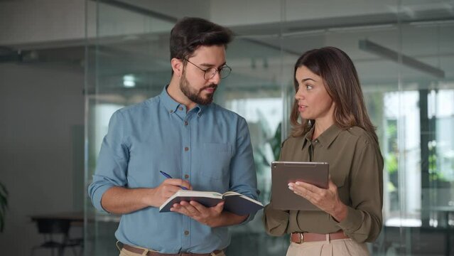 Happy busy latin partners using digital tablet talking standing in office. Professional business people having conversation sharing ideas. Female executive manager working together with male employee.