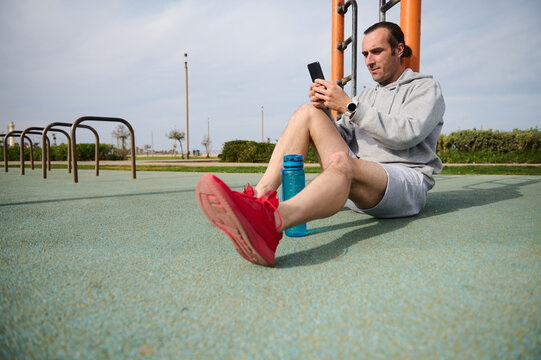 Athletic man checking mobile sports app on his smartphone, analyzing his workout results, sitting on the sportsground