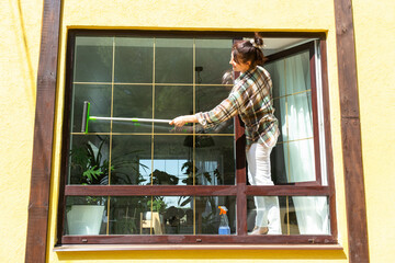 A woman manually washes the window of the house with a rag with a spray cleaner and a mop outside....
