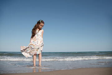 Thoughtful Girl in Dress on Beach Shore. A young girl in a flowing summer dress stands thoughtfully...