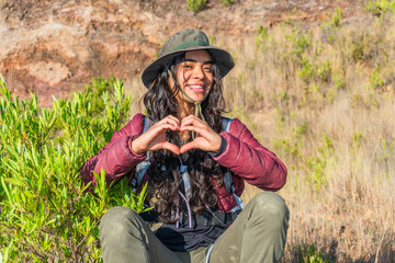 young female traveler with hat sitting on the mountain making the shape of a love heart with her hands. Travel scene