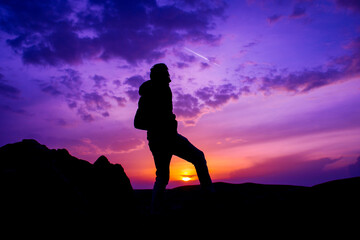A man stands on a hill in front of a purple sky with the sun setting behind him.