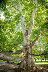 Beautiful woman with gorgeous curly hair doing yoga in nature, dressed in white and yellow combination, sitting on old plane tree. Concept: active, healthy life, in love with nature