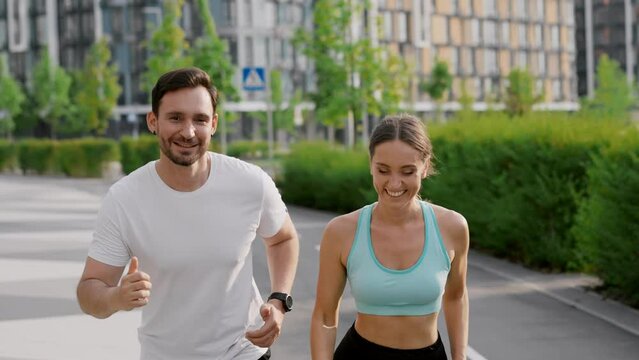 A smiling couple make a high five gesture with touching fists and start morning jogging. A young sportive girl and guy train in the city park area among modern residential highrise buildings. The