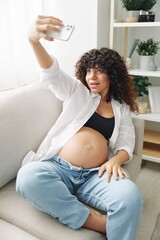 Pregnant woman blogger sits on the couch at home and takes pictures of herself on the phone, selfie and video call, consultation with the doctor online, pregnancy management