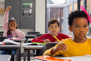 Biracial girl raises her hand in a school classroom; biracial boy in red looks curious