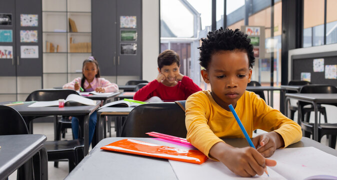 Biracial boy in yellow focuses on writing at his desk in a school classroom