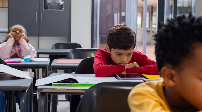 Biracial boy in red looks sad at his desk in a school classroom, with copy space, classmates around 