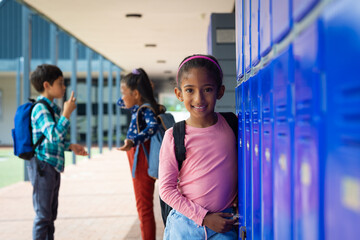 Biracial girl with a pink backpack smiles near school blue lockers, her dark hair tied back