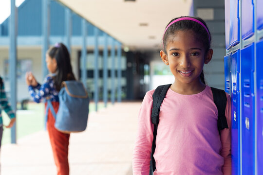 Biracial girl with a pink headband smiles near school blue lockers with copy space, another girl in  - Powered by Adobe