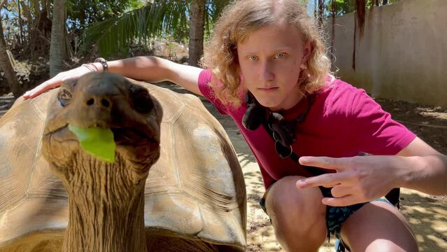 Tourist boy posing for photo with Aldabra giant tortoise endemic species - one of the largest tortoises in the world in the zoo nature park on Mauritius island. Then both noticed it was video shooting