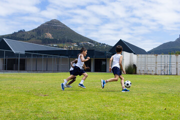 Two boys are playing soccer on a grassy field with mountains in the background in school