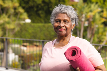 Senior biracial woman with gray hair holds a pink yoga mat outdoors - Powered by Adobe