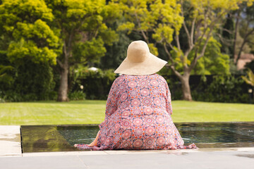 Senior biracial woman sits by a pool, wearing a wide-brimmed hat and a floral dress