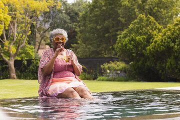 Senior biracial woman enjoys a drink by the poolside, her feet dipped in water, with copy space