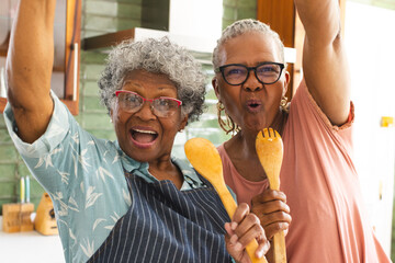 Senior African American woman and senior biracial woman celebrate with kitchen utensils - Powered by Adobe