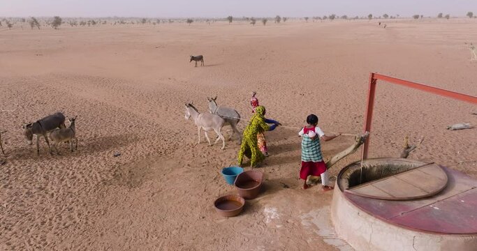 Aerial close-up. Three black woman drawing water from a deep well with the help of donkeys in the barren landscape of the Sahel, Sahara Desert, Senegal. Drought, Climate Change, Desertification