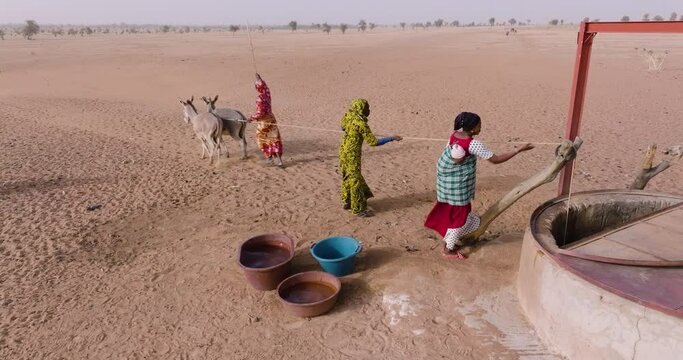 Aerial close-up. Three black woman drawing water from a deep well with the help of donkeys in the barren landscape of the Sahel, Sahara Desert, Senegal. Drought, Climate Change, Desertification