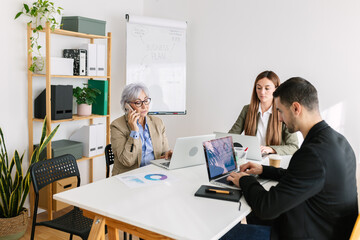 Diverse group of professional coworkers sitting in the same table in a meeting room using laptop and mobile phone. Teamwork and business concept.