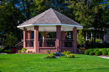 Fototapeta na wymiar A large multi-sided red brick gazebo with black metal railing. The round roof is made of brown asphalt shingles. There's a lush green lawn, shrubs and flowers surrounding the building. 