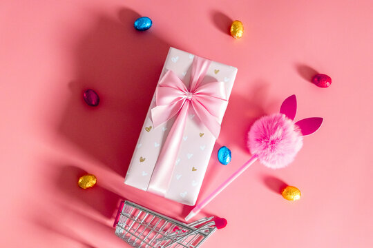 Gift box with bow, bunny handle, mini trolley and Easter eggs on a pink background.