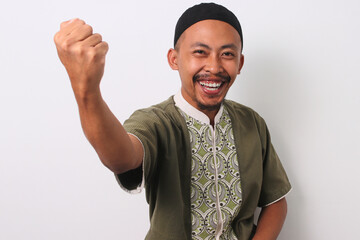 A joyful Indonesian Muslim man in koko and peci raises his arm and smiles in celebration of a...