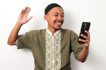 A happy Indonesian Muslim man in koko shirt and peci video calls with his family in his hometown,...