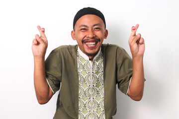 Indonesian Muslim man in koko shirt and peci celebrates with crossed fingers, expressing joy and...