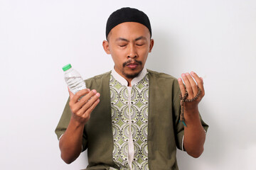 An Indonesian Muslim man in koko and peci raises his hands in prayer while holding a bottle of...