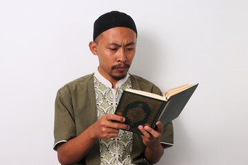 An Indonesian Muslim man reciting the Holy Quran with focus during Ramadan. Isolated on a white...